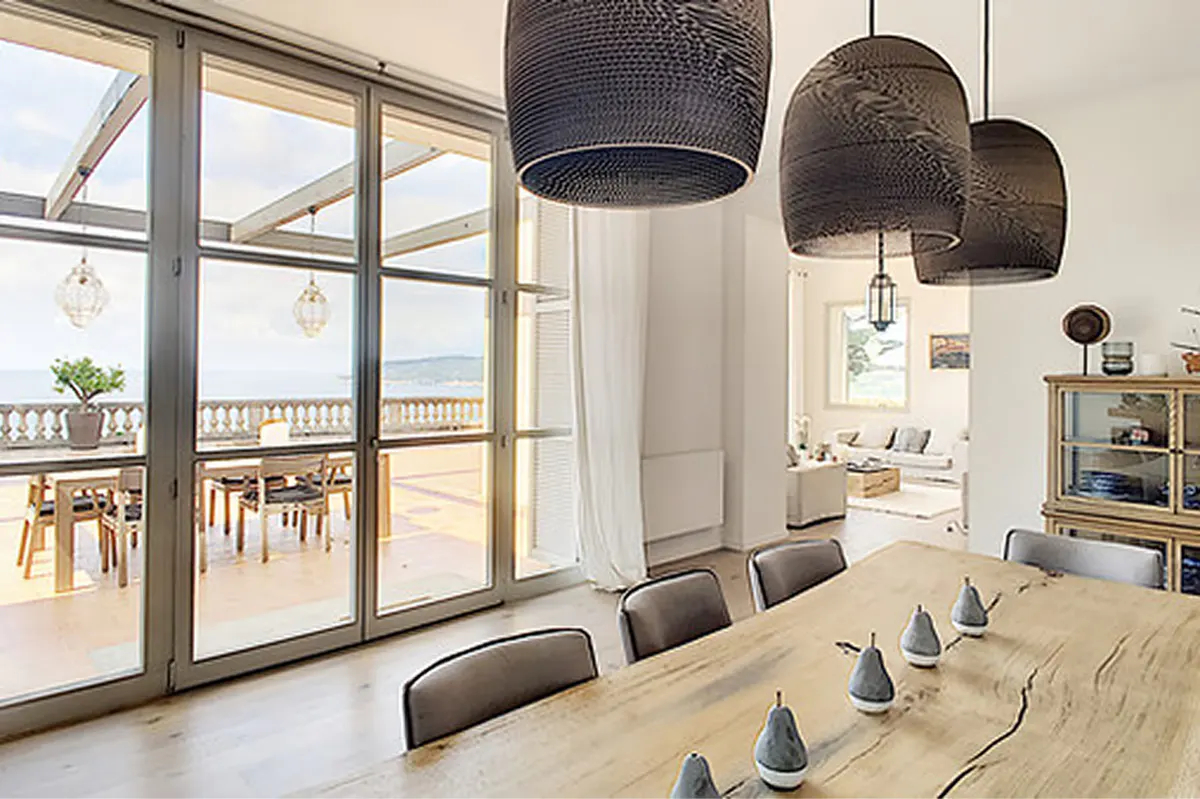 Dining room with beige walls, wooden table, and pendant light fixtures, coastal views beyond
