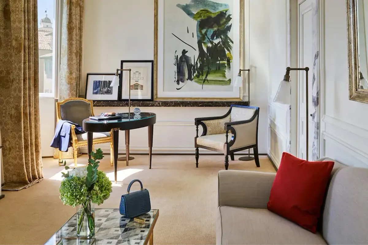 A suite with a desk, sitting area with red accent pillows, artwork, and tall windows with gold drapery