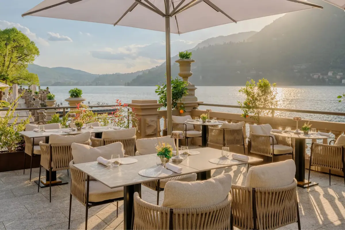Covered terrace with round tables and chairs positioned along waterfront with mountains beyond