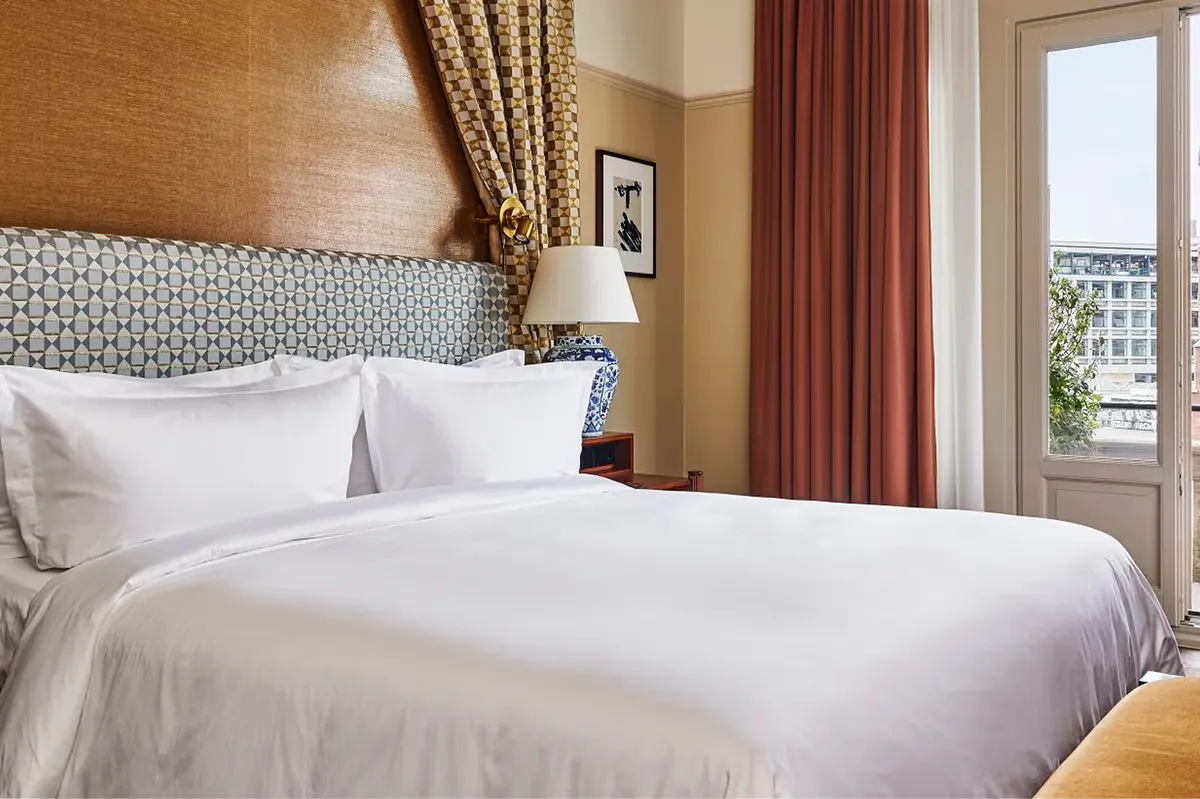 Bed with neutral linens, patterned headboard, and river view through tall windows