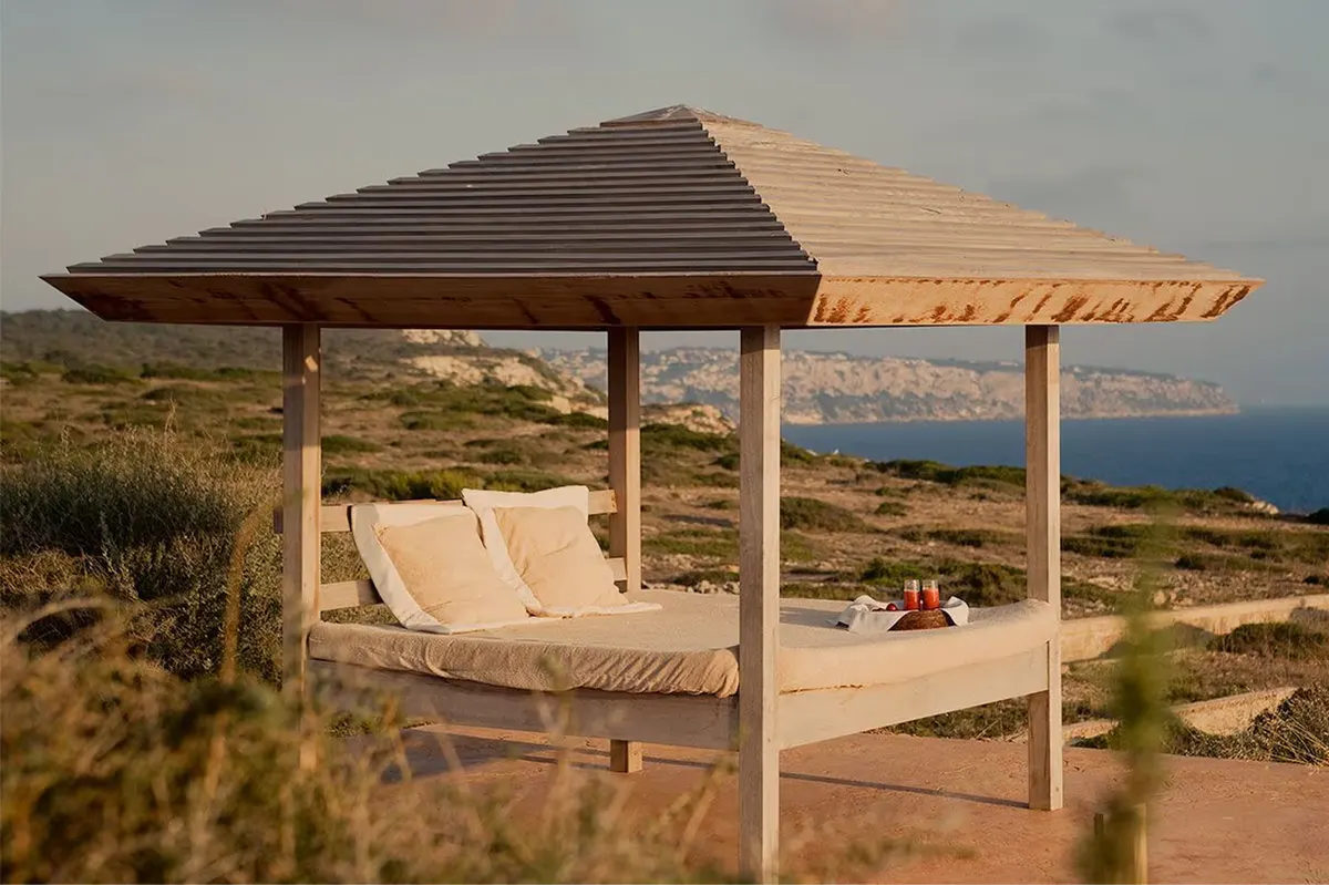 A bed with pale curtains and wooden ceiling, framed by an open terrace and coastal light
