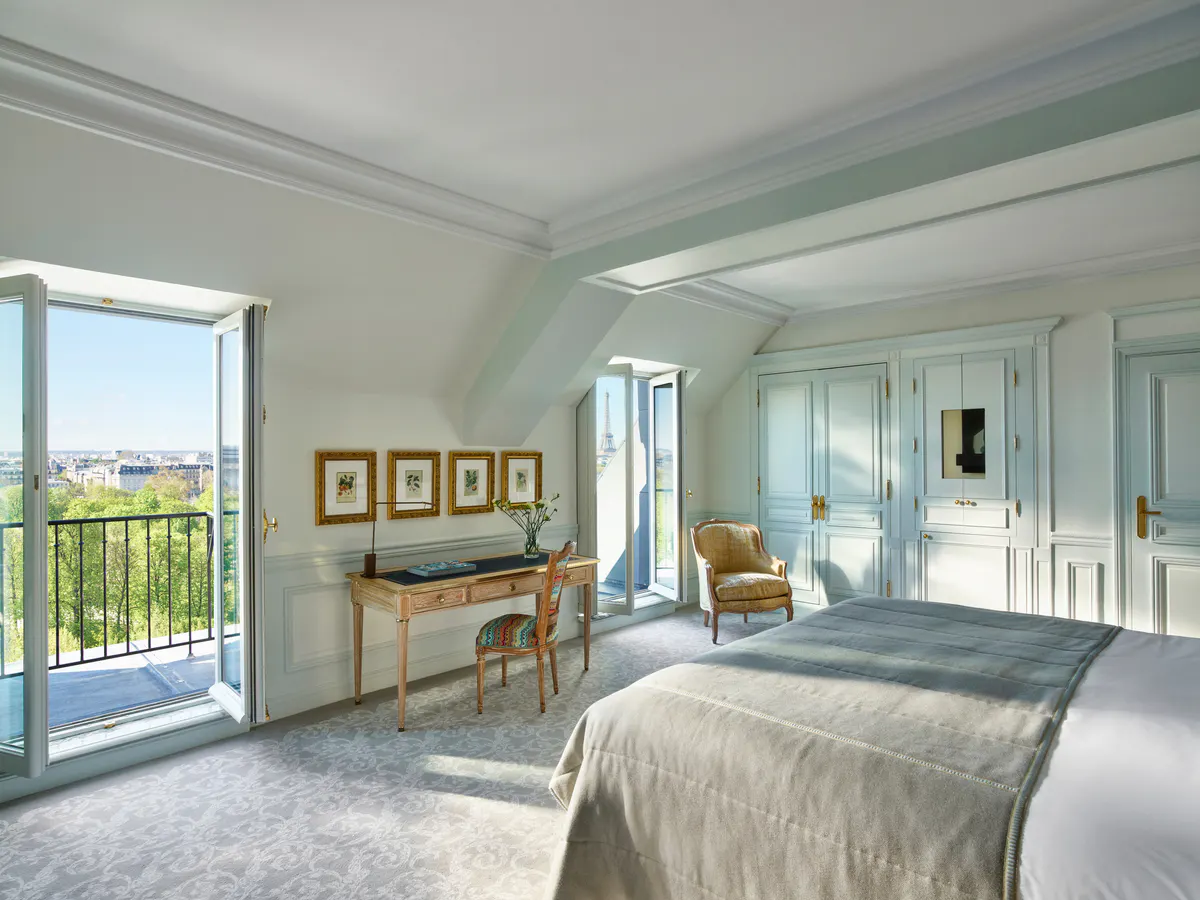 A bedroom with pale walls, queen bed, and doors opening onto a blue-railed Parisian balcony