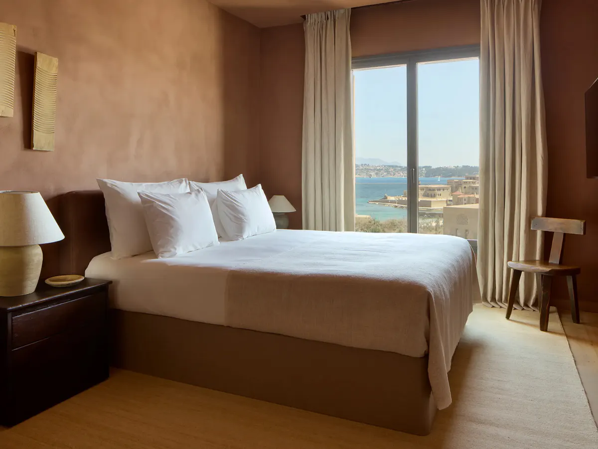 Pillows are piled high on a bed facing arched windows over the sea and stone ramparts
