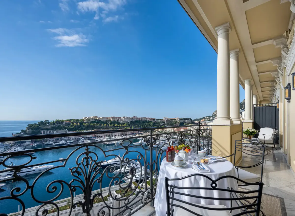 Wrought-iron terrace railing with sea and coastal buildings visible under blue sky