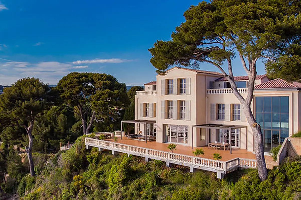 Pale stone manor on cliff overlooking bright blue Mediterranean waters and tree-covered slopes