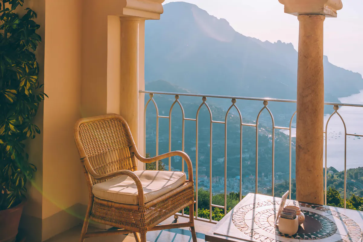 Wicker lounge chair on cream terrace with mountains, sea, and potted plants in golden light