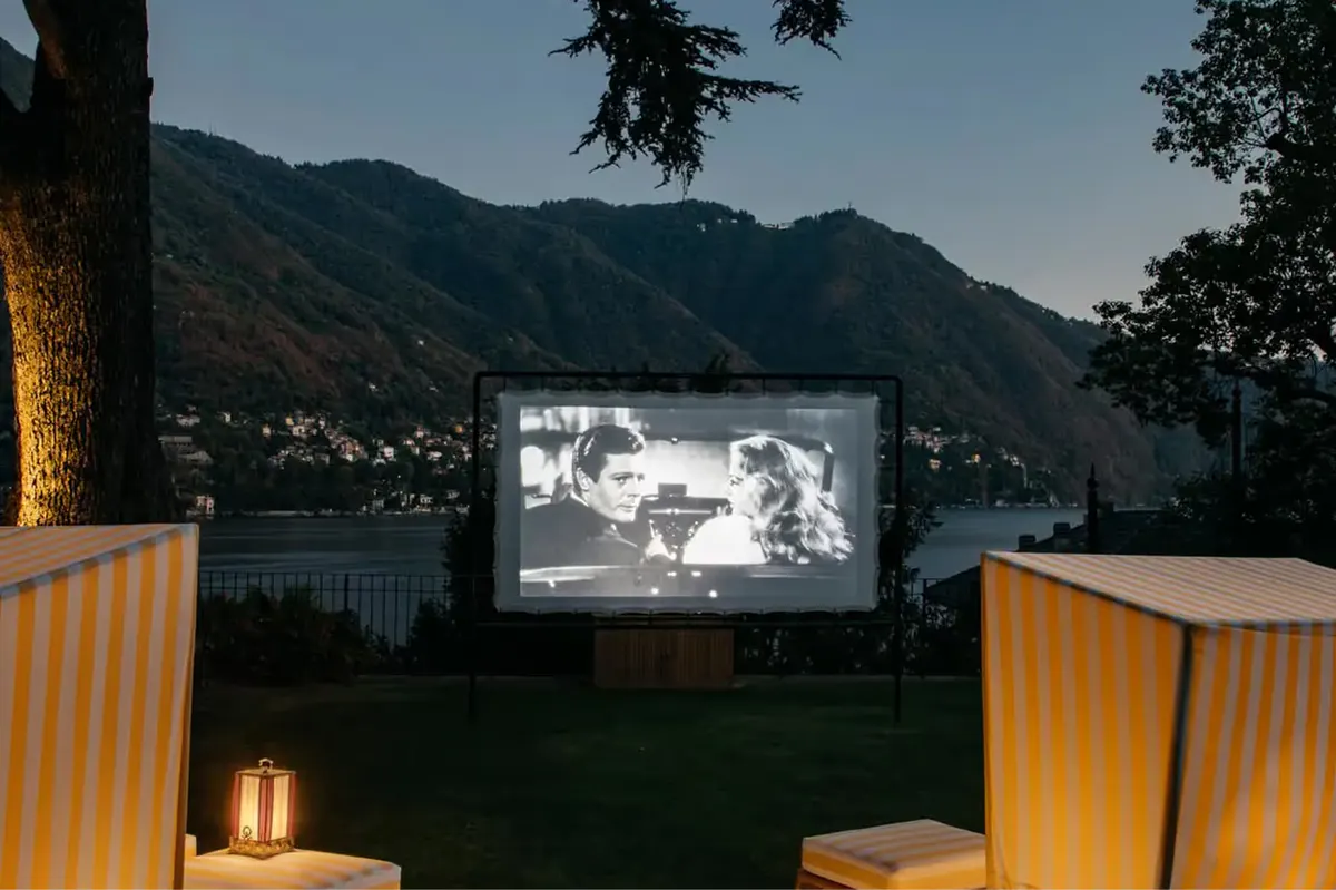 An outdoor movie screen glowing at dusk with upholstered seating and lake view