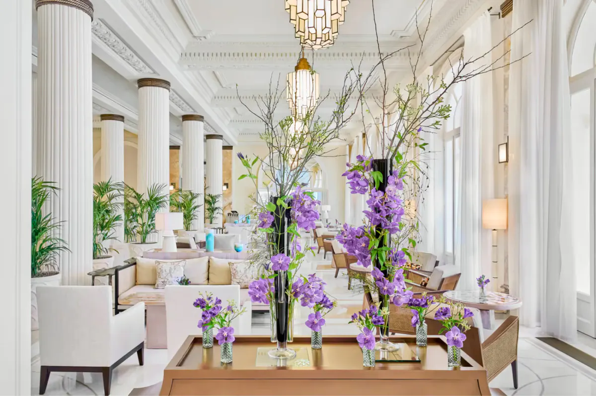 Bright lobby with tall white columns, purple flower arrangements on a wooden table, and chandelier lighting