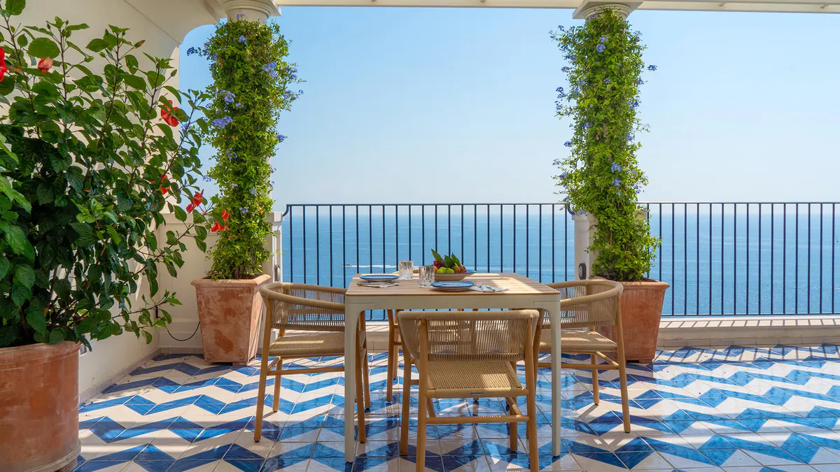 Outdoor terrace table with wooden chairs overlooking sea, ivy-draped pergola frame