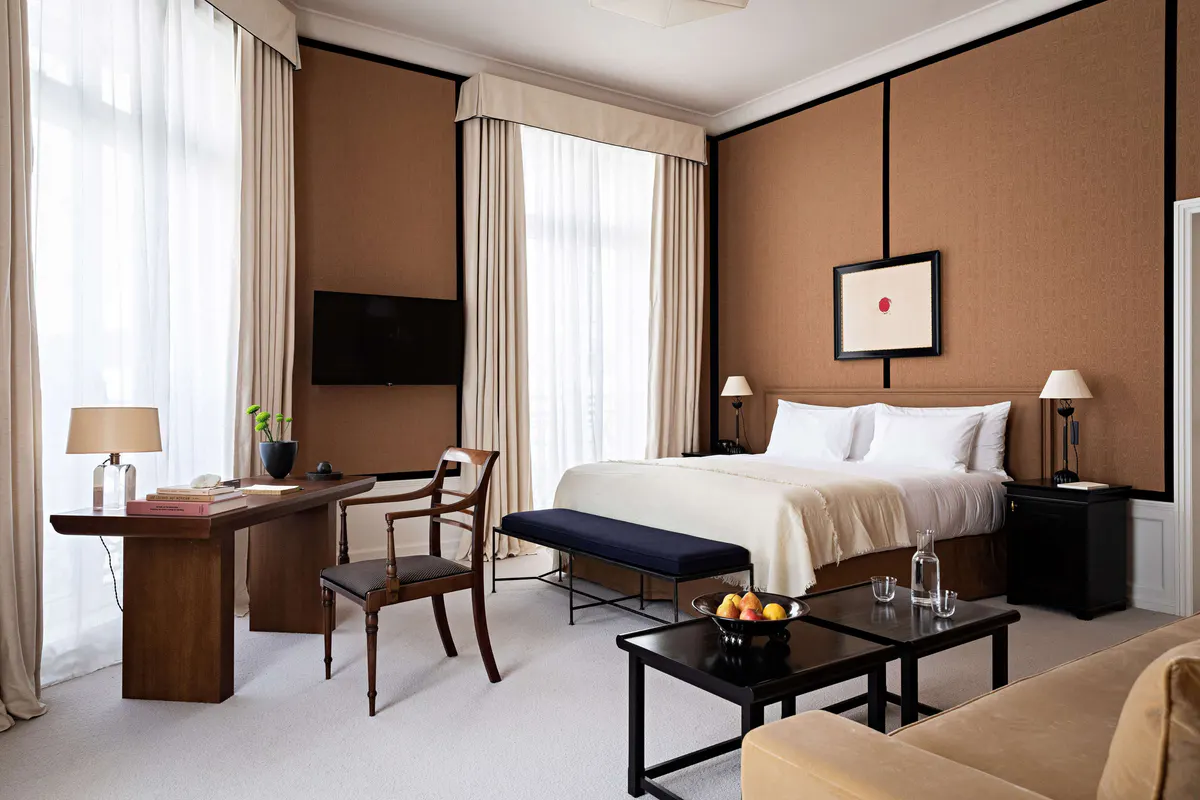 A bedroom with tan and brown tones, wood furnishings, and tall windows