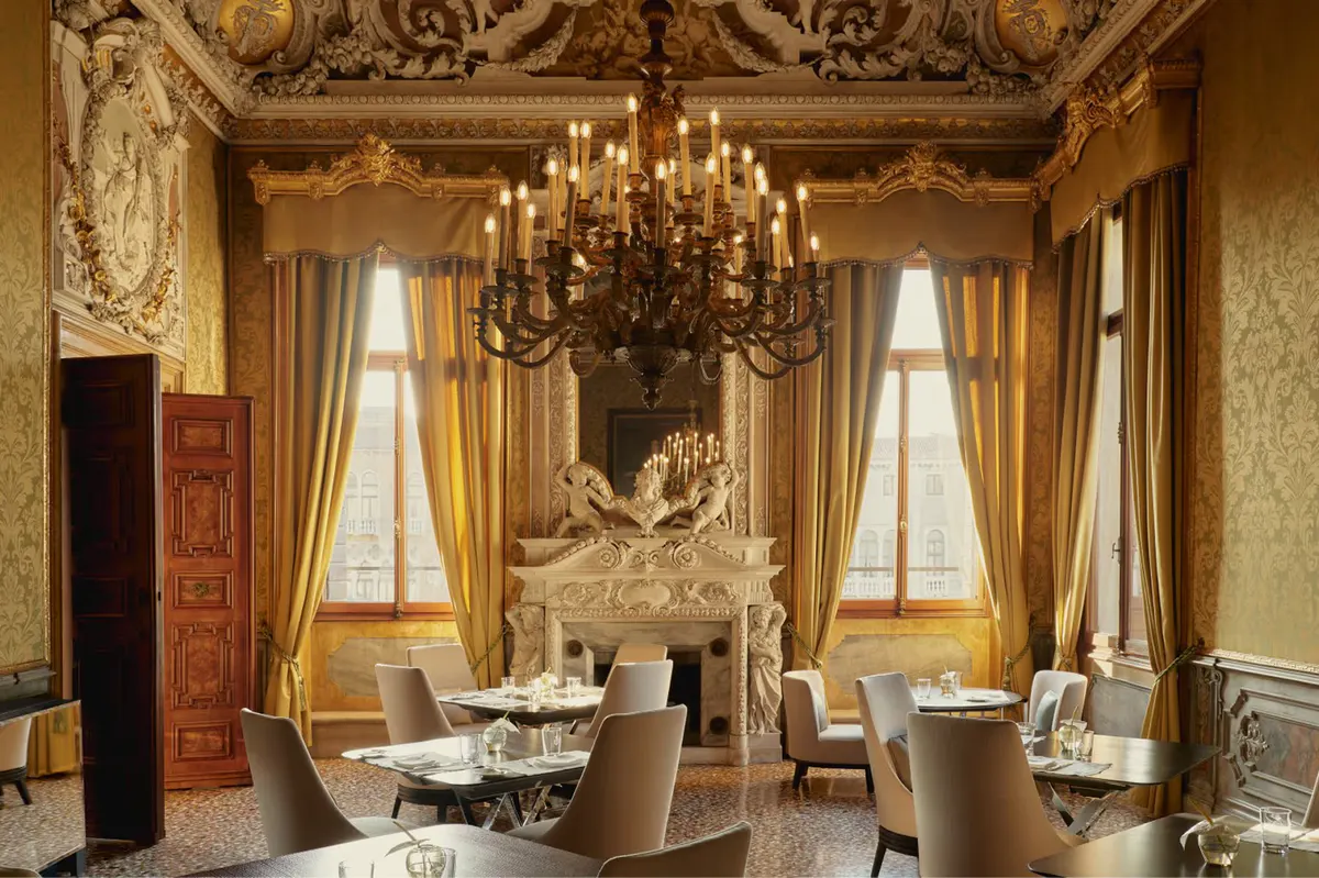 A formal dining room with ornate gilded walls, crystal chandelier, draped windows, and round tables with chairs