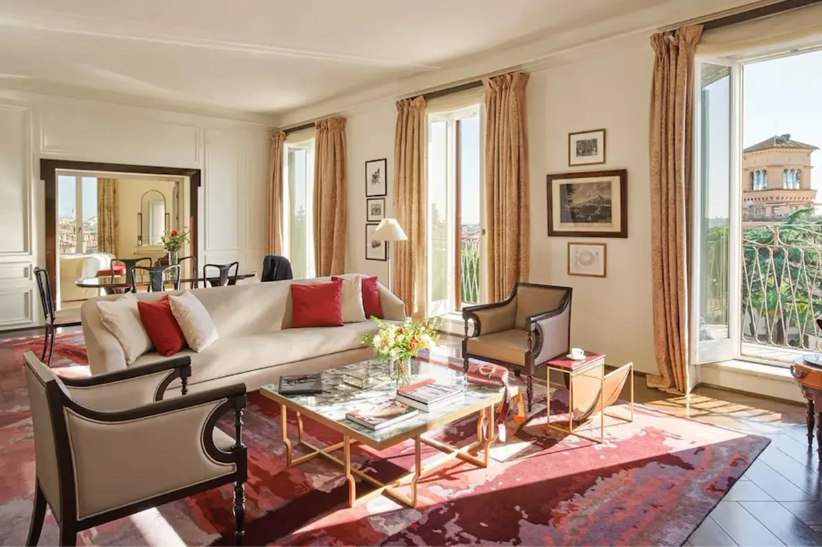 A living room with cream furnishings, red accent chairs, decorative artwork, and tall windows overlooking Rome