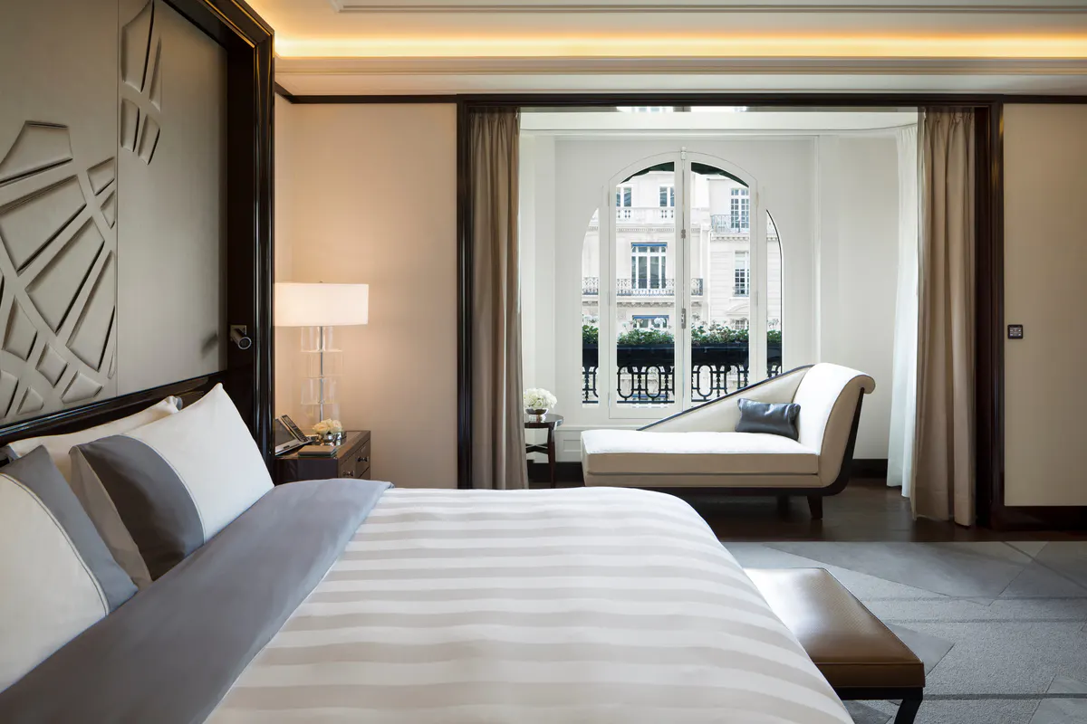 Hotel suite with bed facing a window, warm lighting, wall sconces, and period furnishings