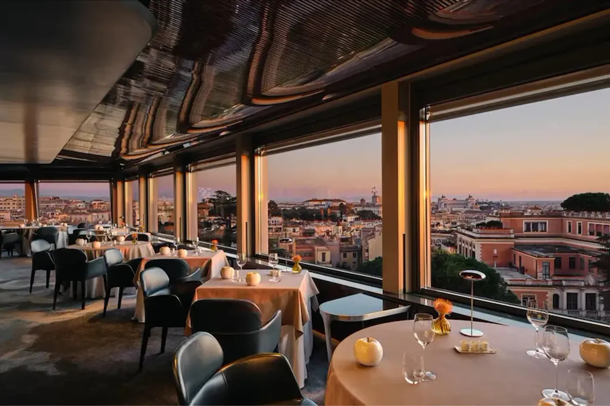 La Terrazza rooftop restaurant at dusk with floor-to-ceiling windows, diners at round tables, Rome’s skyline in the background