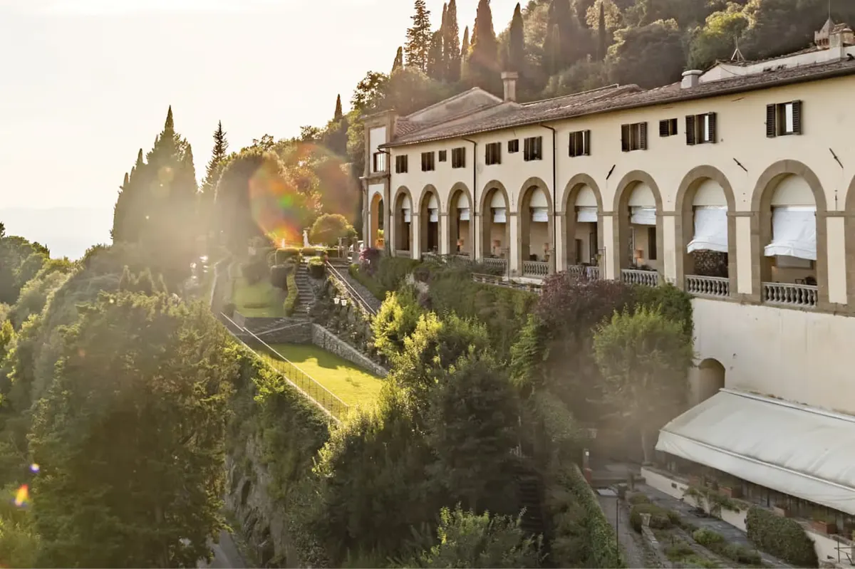 Cream building with arched columns, shuttered windows, cypress trees, and golden light across the hillside