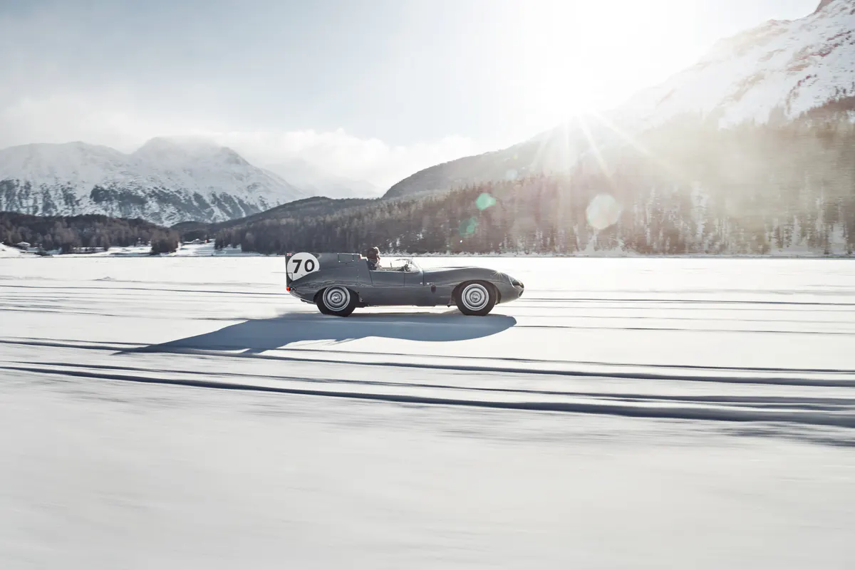 A vintage car racing on a snowy alpine lake with forested mountains beyond