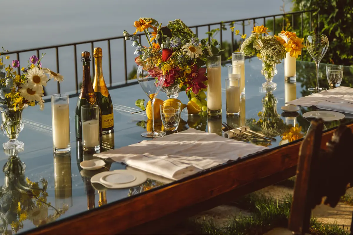 Cliffside table setting with floral arrangements, golden wine glasses, sea vista and mountains beyond