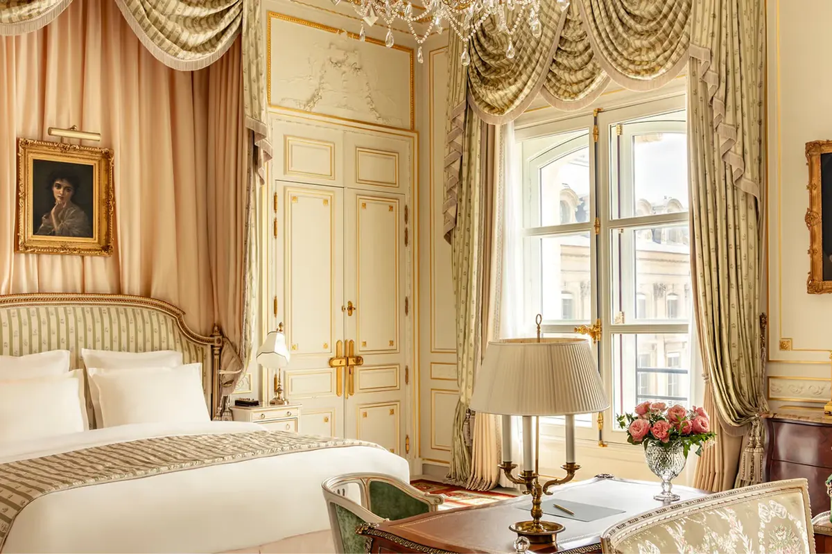 A bedroom with draped gold curtains, gilt cove ceiling, damask wallpaper, and cream bedding