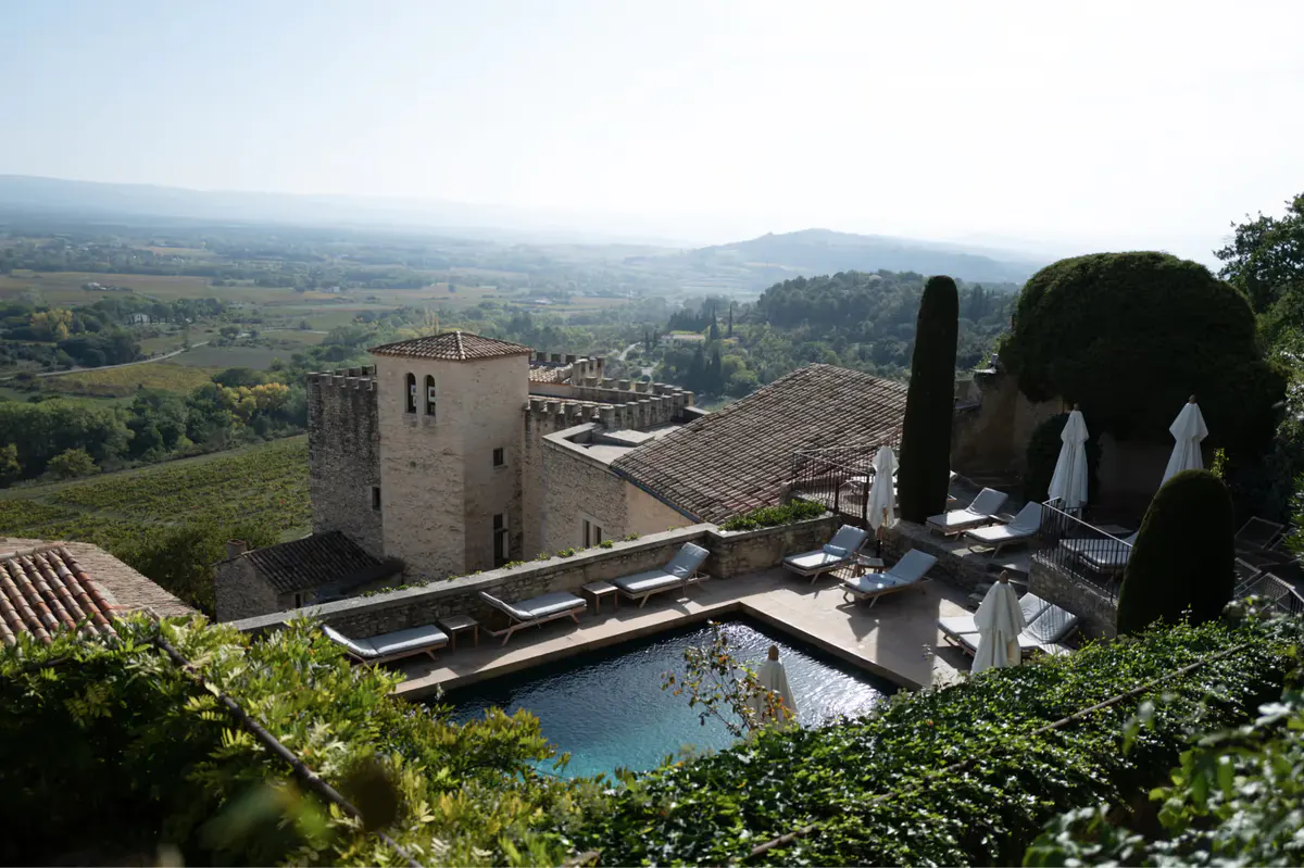 Medieval stone buildings with turquoise pool, sun loungers under white umbrellas, boxwood hedges, and far countryside