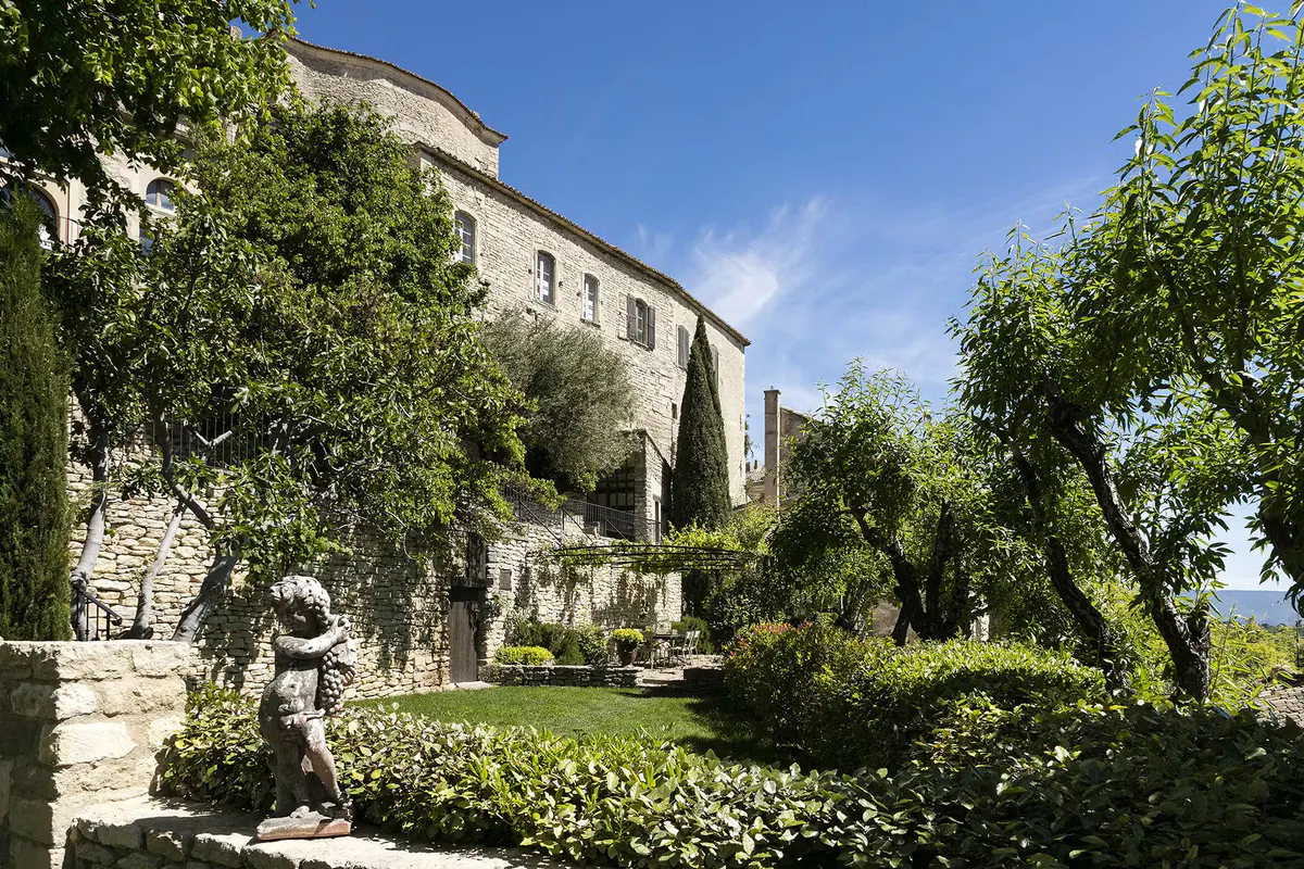 Hotel exterior with stone walls, cypress trees, and manicured hedges in afternoon sun
