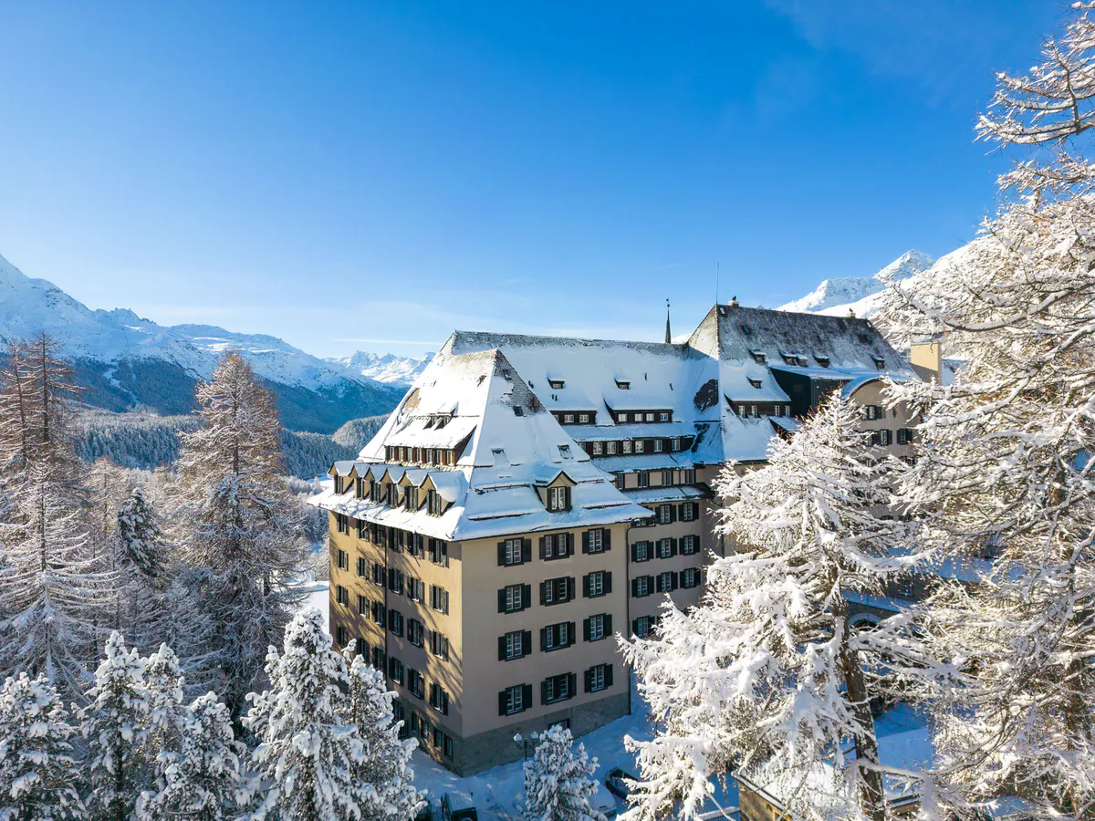 A white Belle Époque hotel on a forested mountain plateau with snow-capped peaks