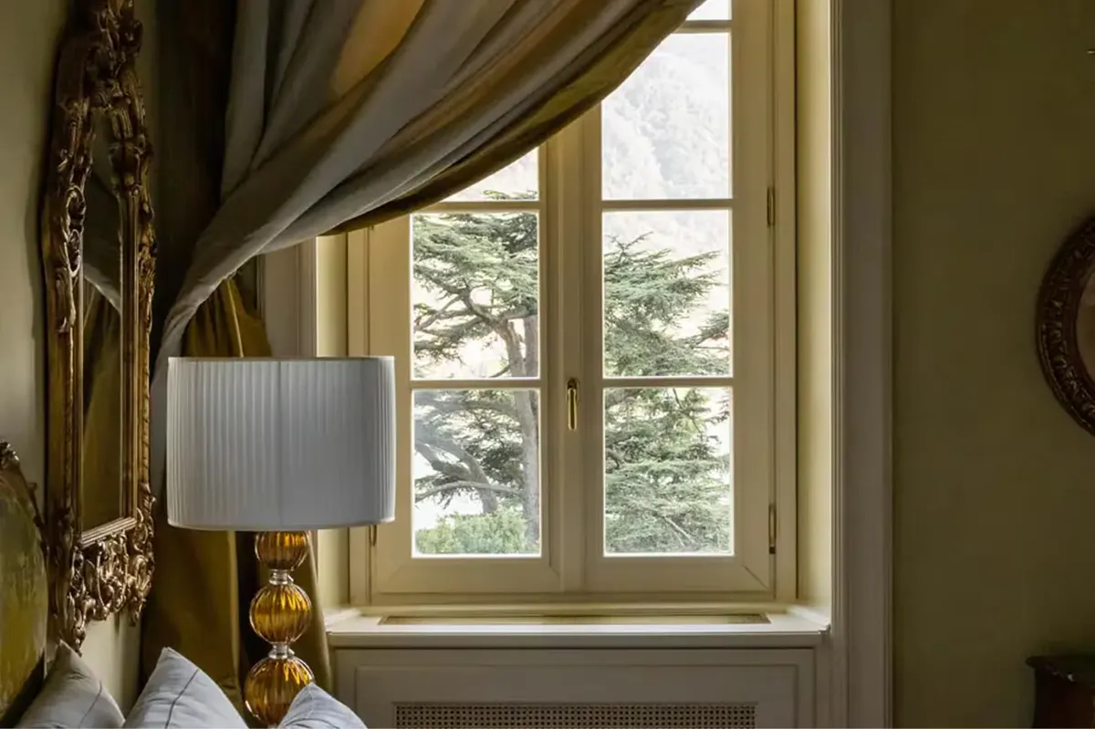 A window with gold damask curtains, lamp, and lake landscape visible through panes
