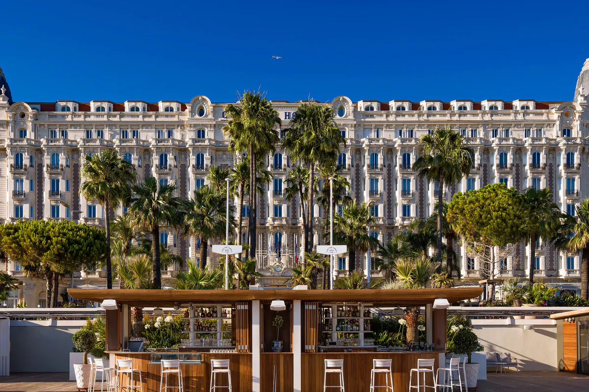 Front facade of Carlton Cannes overlooking the Mediterranean with palm-lined beach and turquoise water