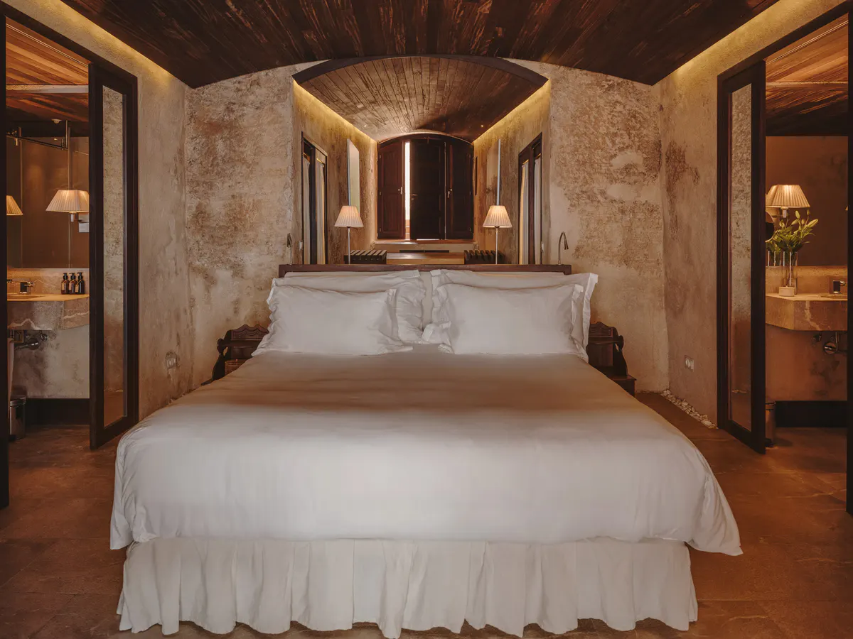 A spacious bedroom with a white bed, wooden headboard, pale ochre walls, and wooden ceiling beams