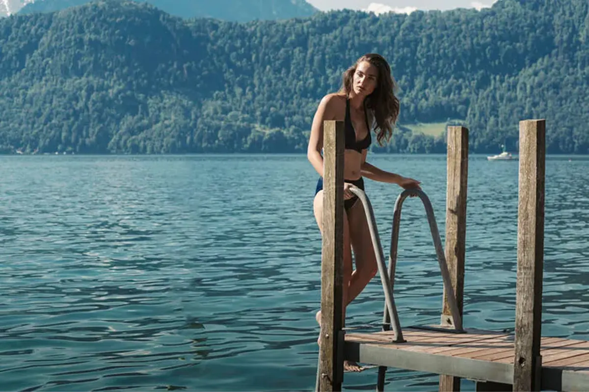 Woman at lake dock in tan swimwear, Lake Lucerne, Alpine mountains behind on clear day
