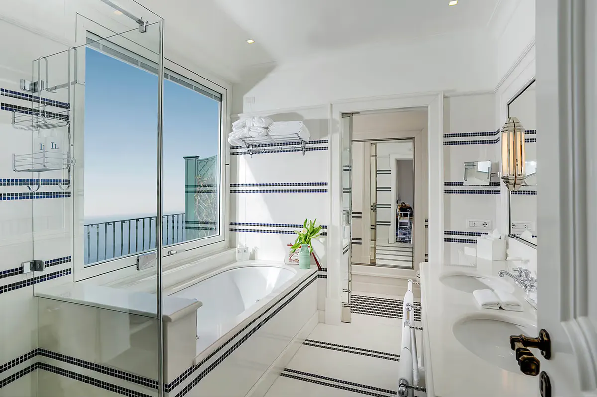 White bathroom with glass shower enclosure, spa tub, marble accents, and view of sea beyond