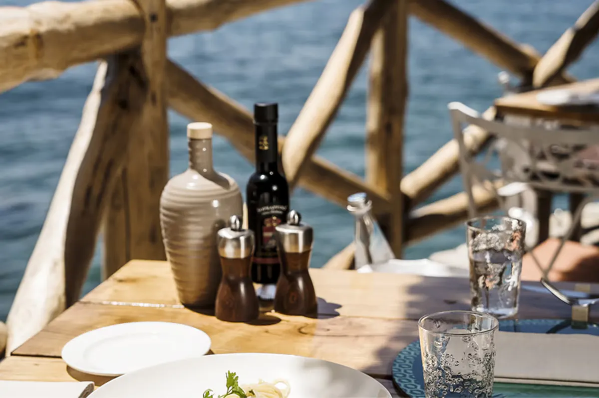 Place setting on wooden table with wine bottles, glasses, and driftwood railing overlooking sea