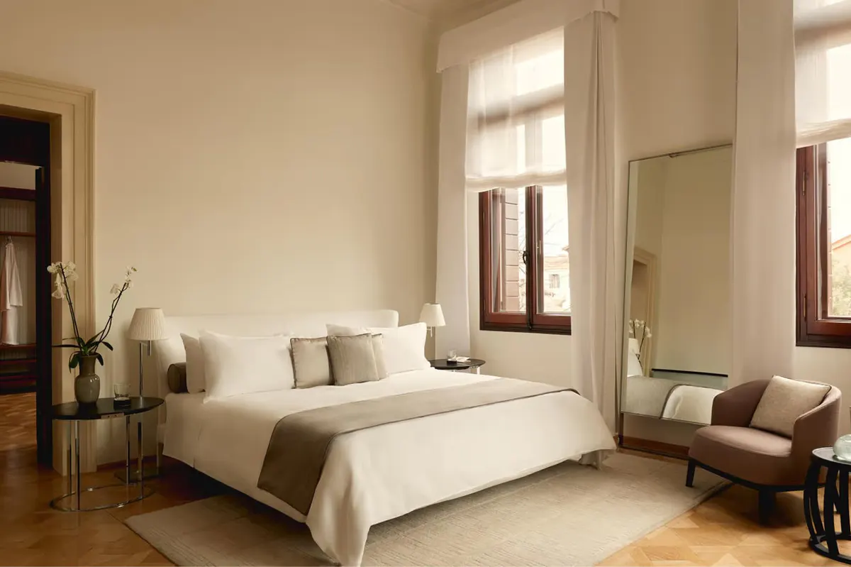 A bright bedroom with neutral linens, brown leather seating, tall windows with natural light, and wooden flooring