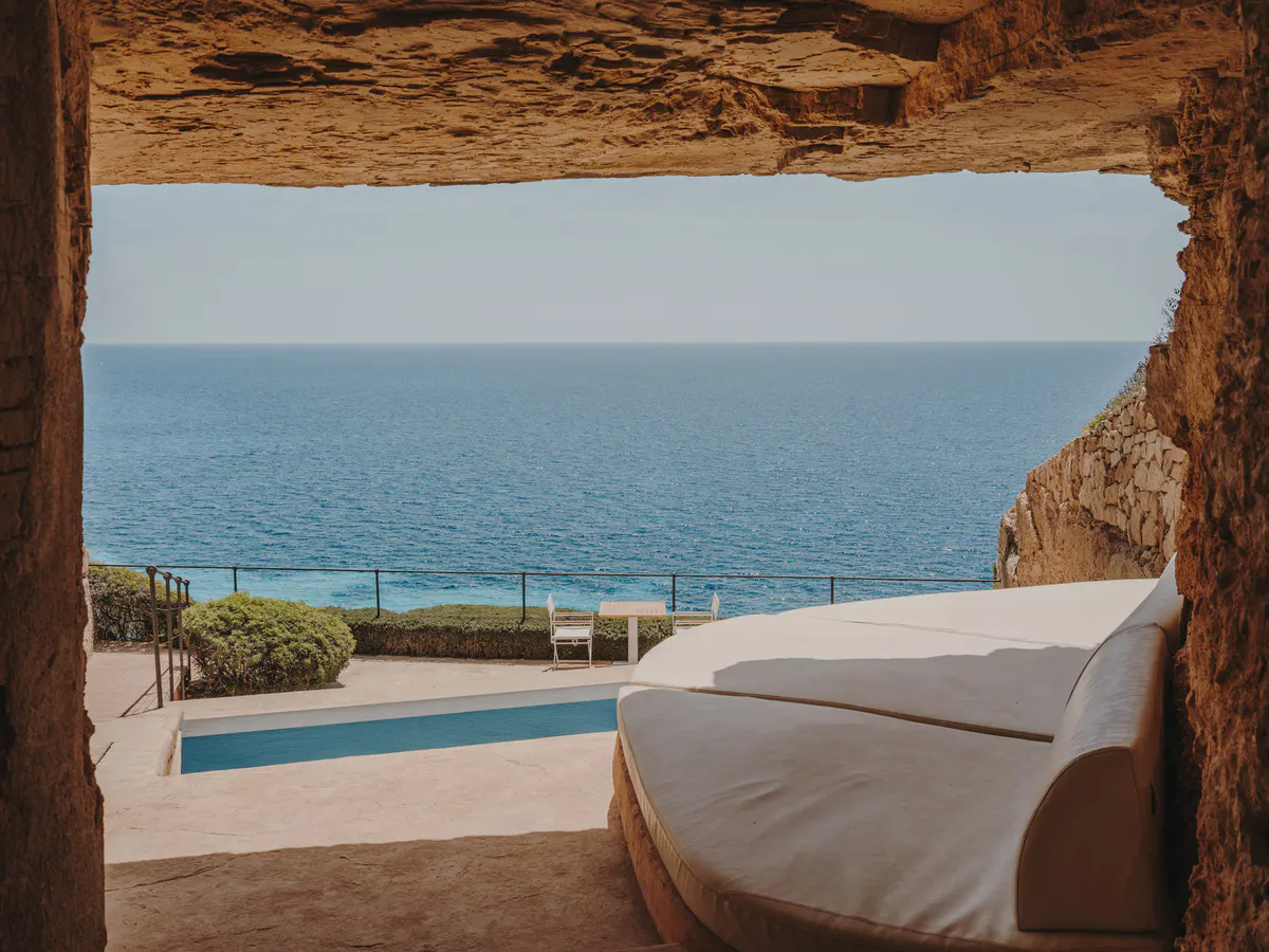 A figure in a tan linen coat walks beside a turquoise pool under sun and coastal cliffs
