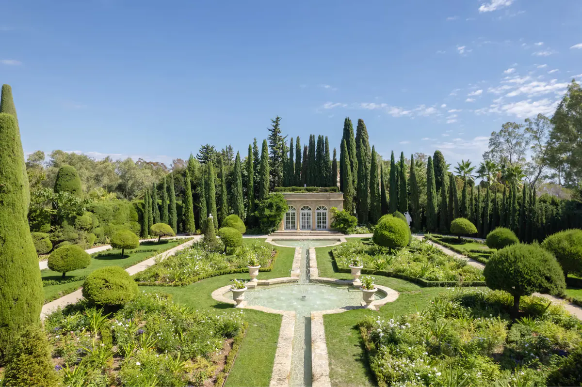 Formal gardens viewed from above, with stone pathways, cypress trees, and château roofline