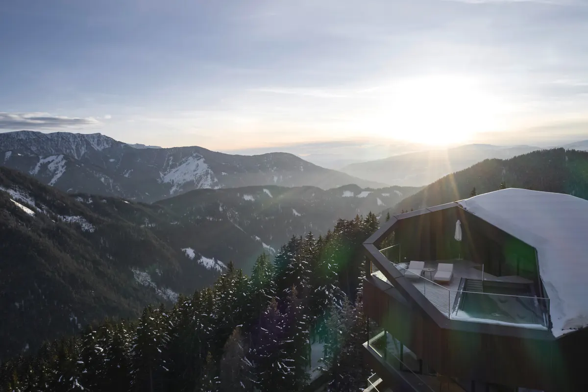 An aerial view of the building’s modern architecture set in mountains at sunset with forested slopes