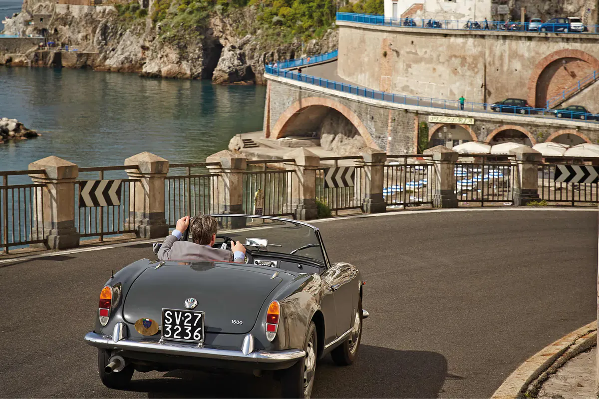 Vintage convertible car parked on coastal bridge with waterfront buildings and boats