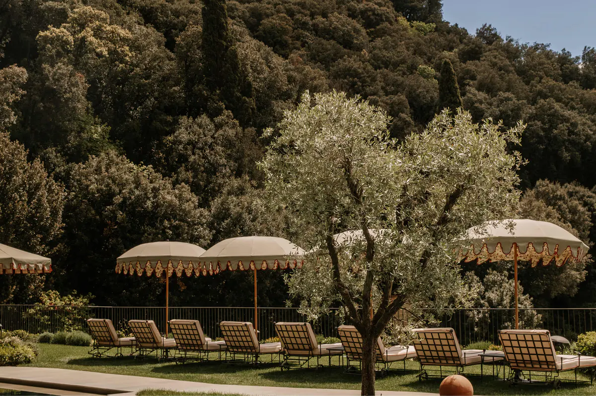 Lounge chairs beneath striped umbrellas in a shaded garden with olive trees and grounds