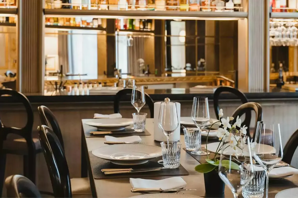 A bar interior with lit shelving behind counter, formal place settings, and glass tabletop