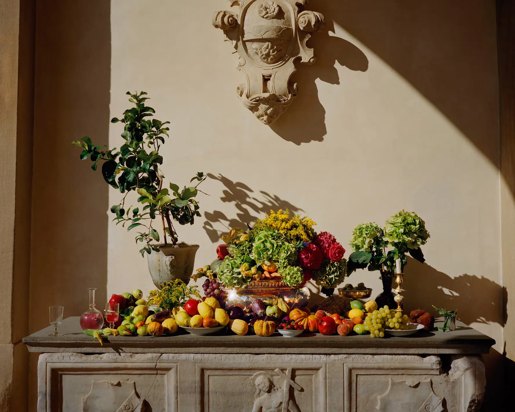 A still life of Tuscan fruit, vegetables and flowers arranged along a carved stone console in raking light
