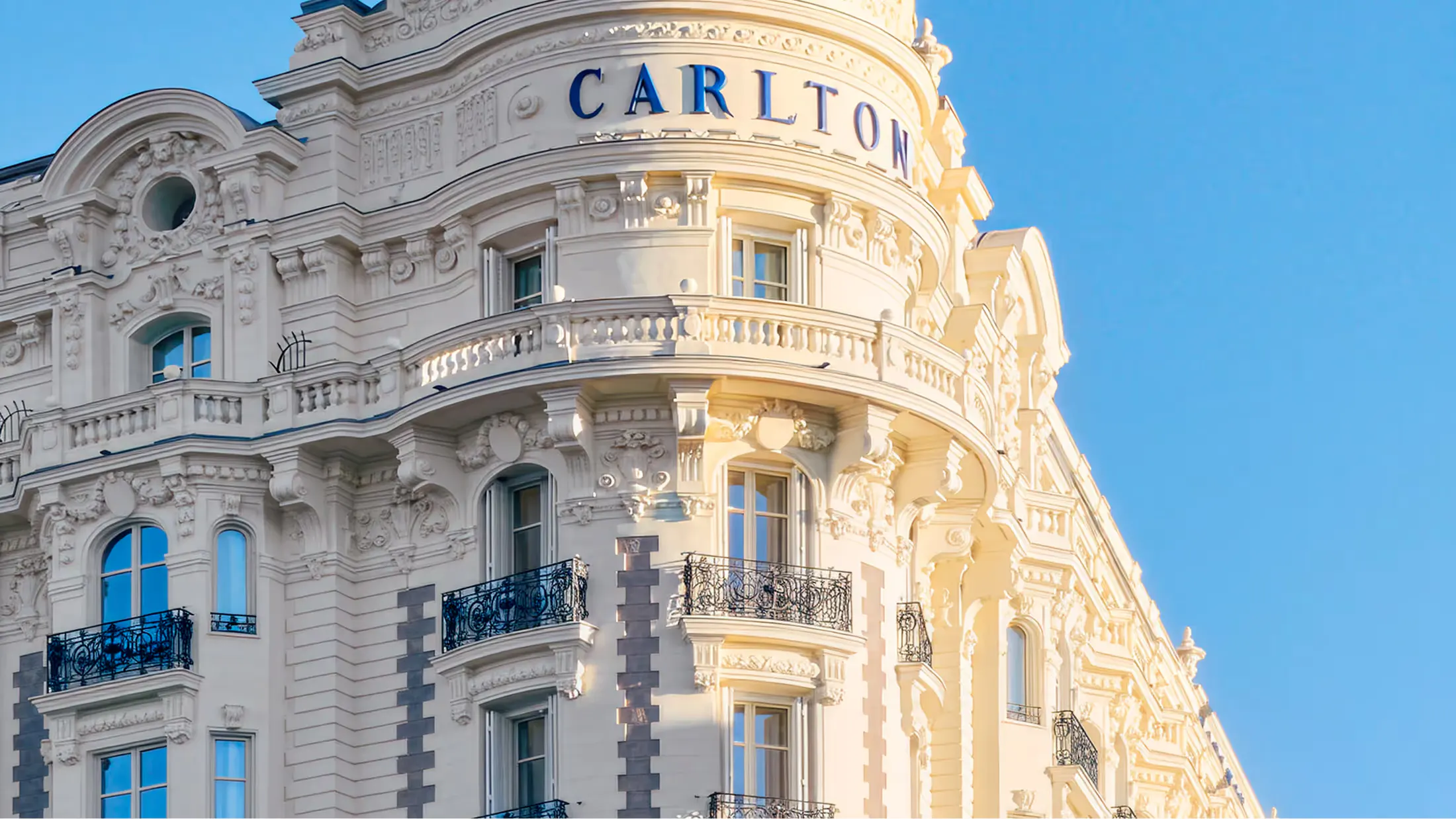 The curved cream stone facade of Carlton Cannes with its iconic turret dome under clear blue sky