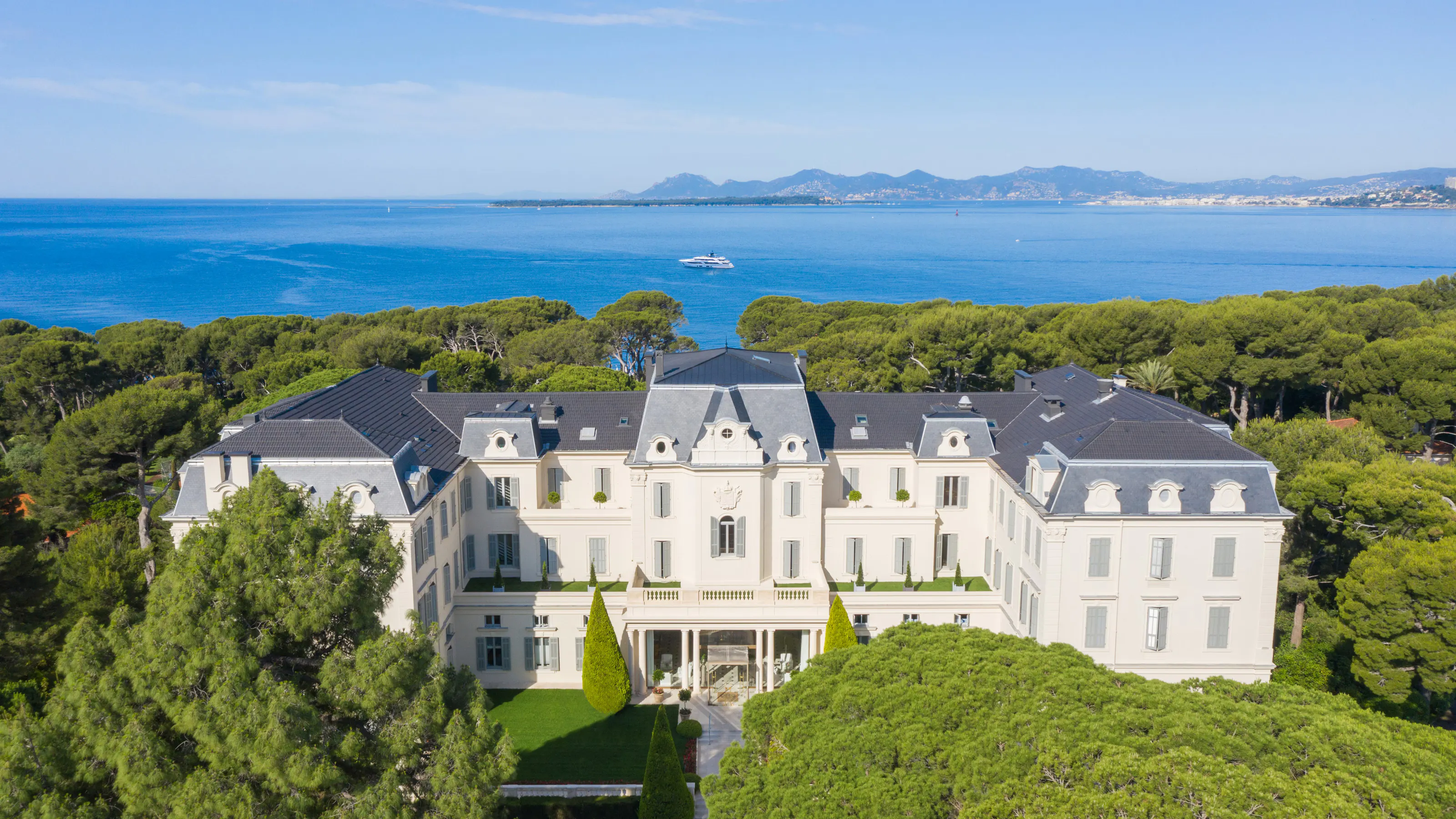 A sweeping aerial view of a whitewashed stone mansion with manicured grounds, pine trees, and the Mediterranean coastline in the background