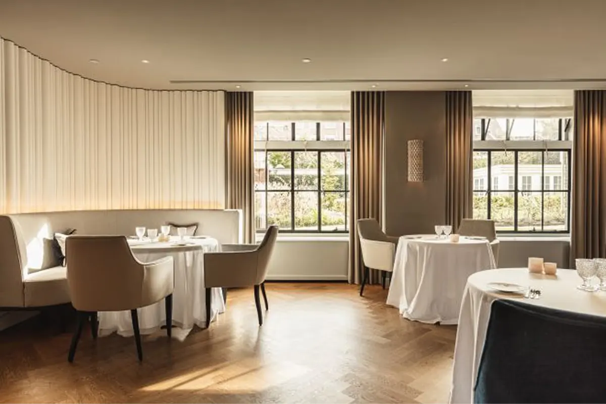 A formal dining room with cream curtains, natural light from tall windows, and white linens on spaced tables