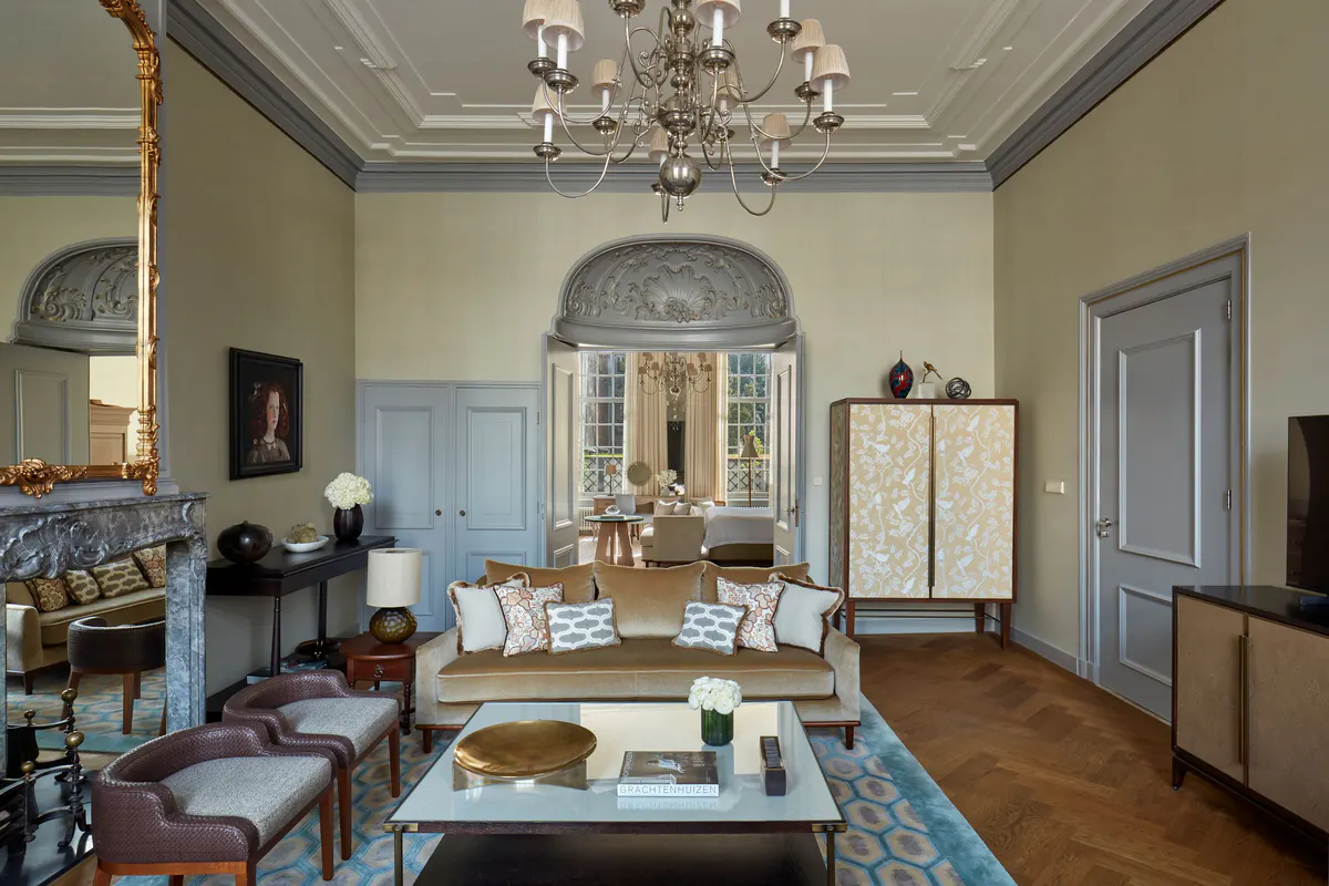 A formal sitting room with muted green walls, gold-framed artworks, and blue cushioned seating arranged around a marble fireplace