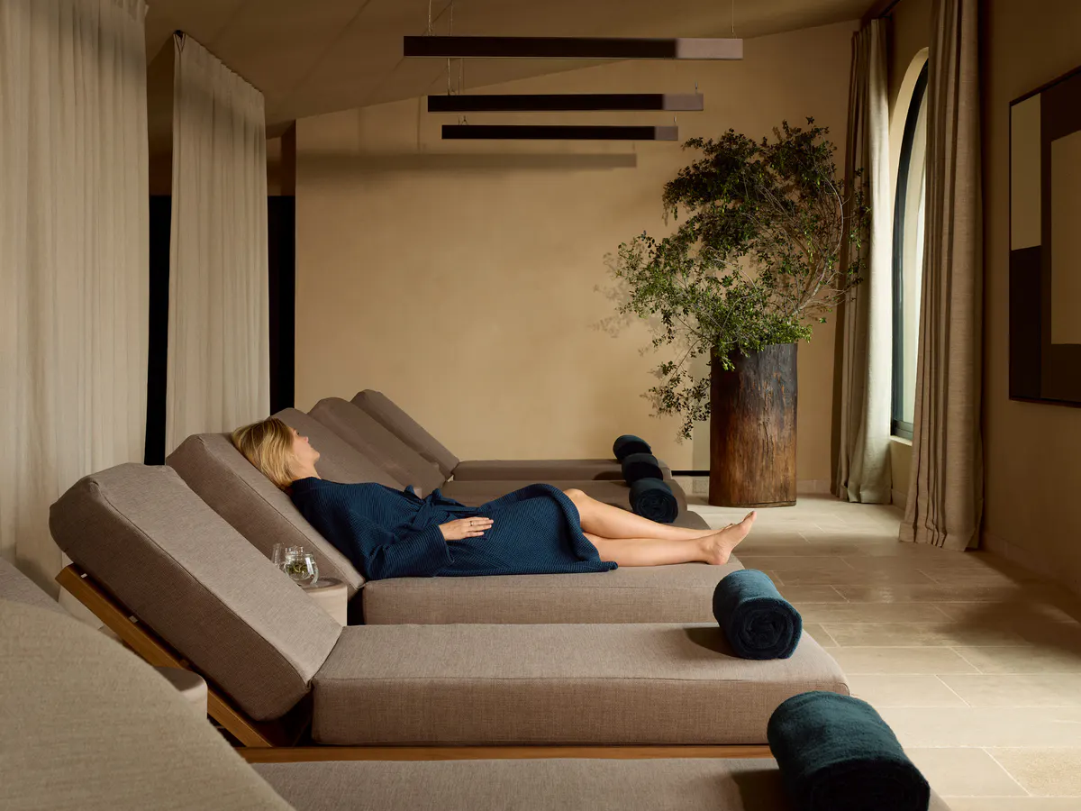 A guest in a robe reclines on a spa lounger beneath pendant lights and a potted olive tree
