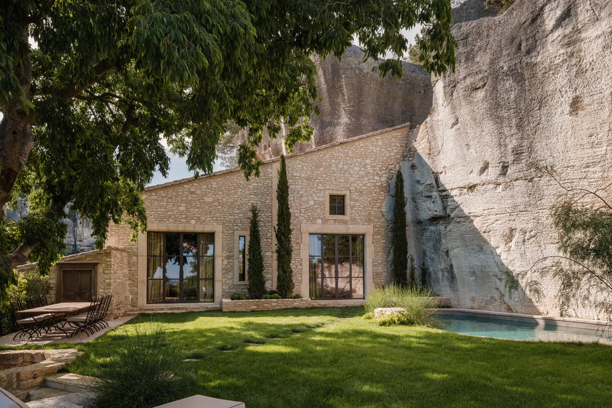 Pale stone villa facade with modern glass doors, cypress plantings, turquoise pool, limestone cliff wall