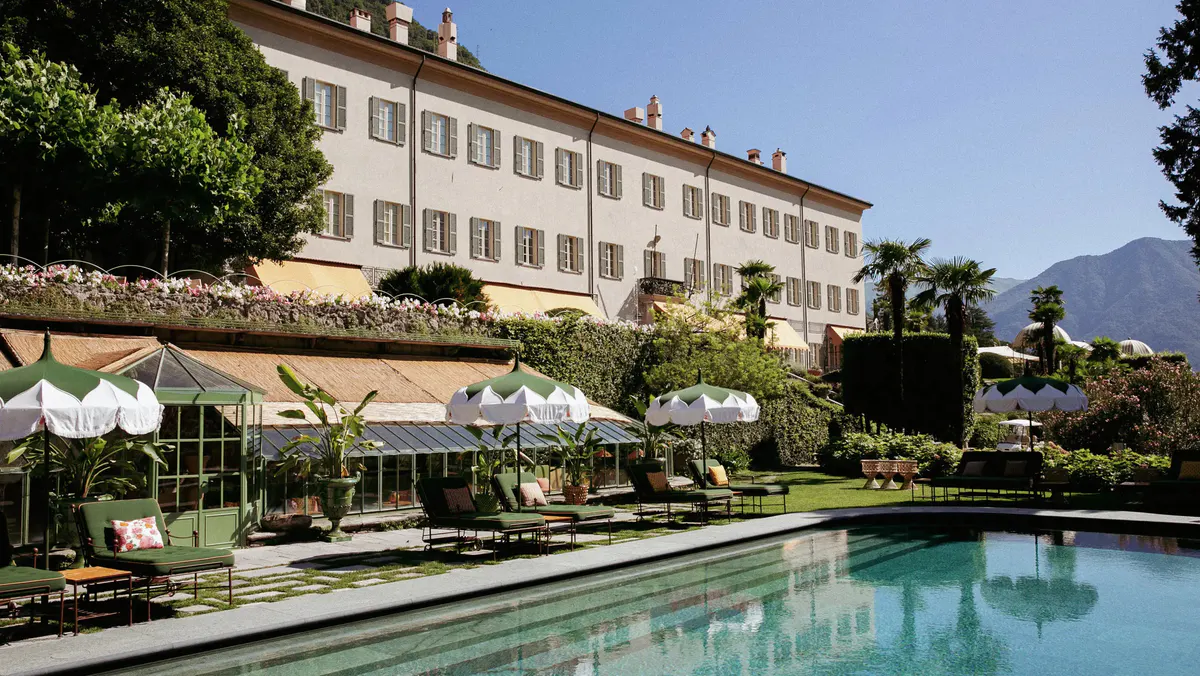 A lakeside palazzo facade with pool terrace and white umbrellas, mountains behind