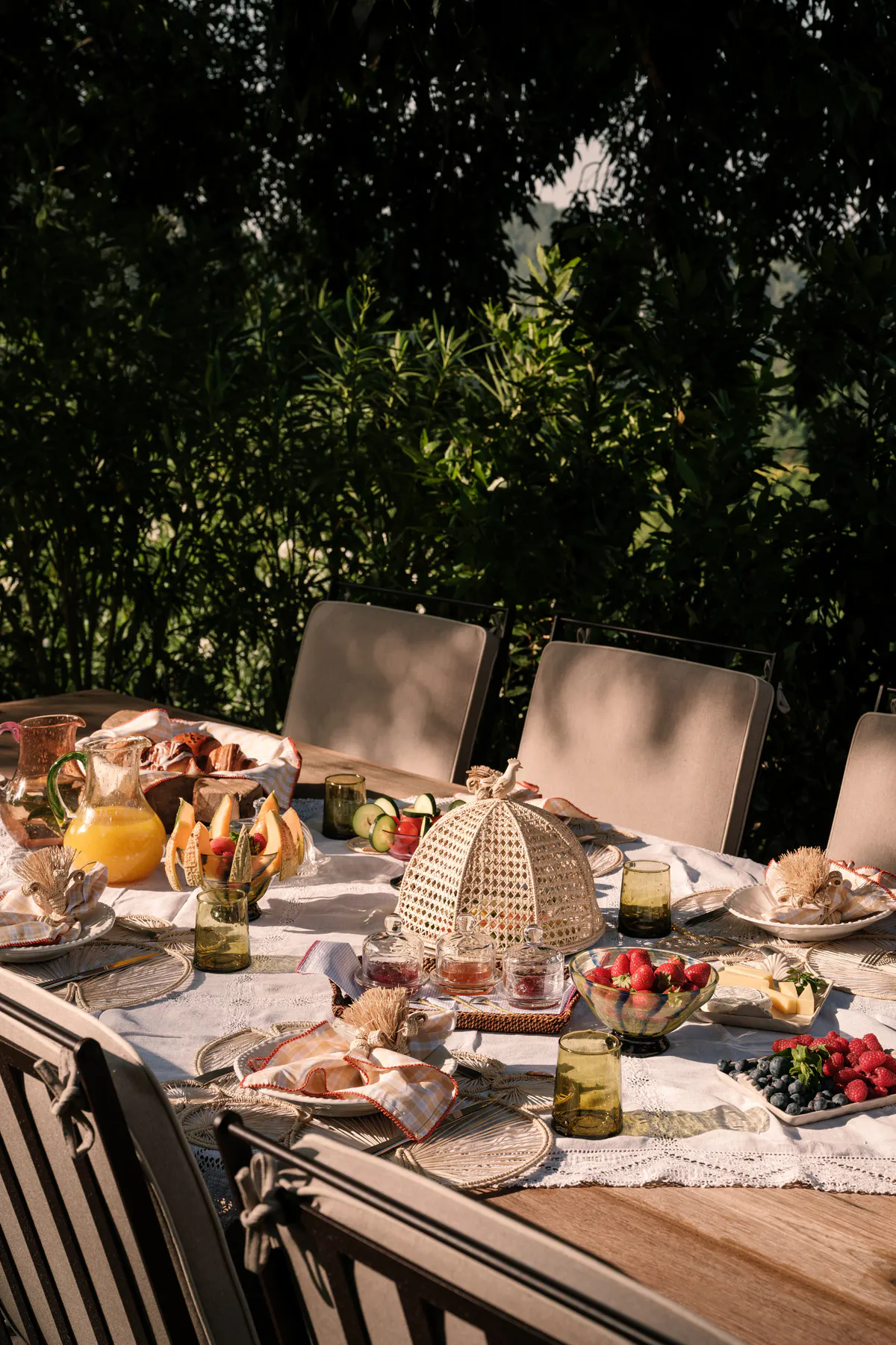 Outdoor breakfast table set with pastries and fruit beneath woven shade sail, lush green foliage, dappled light