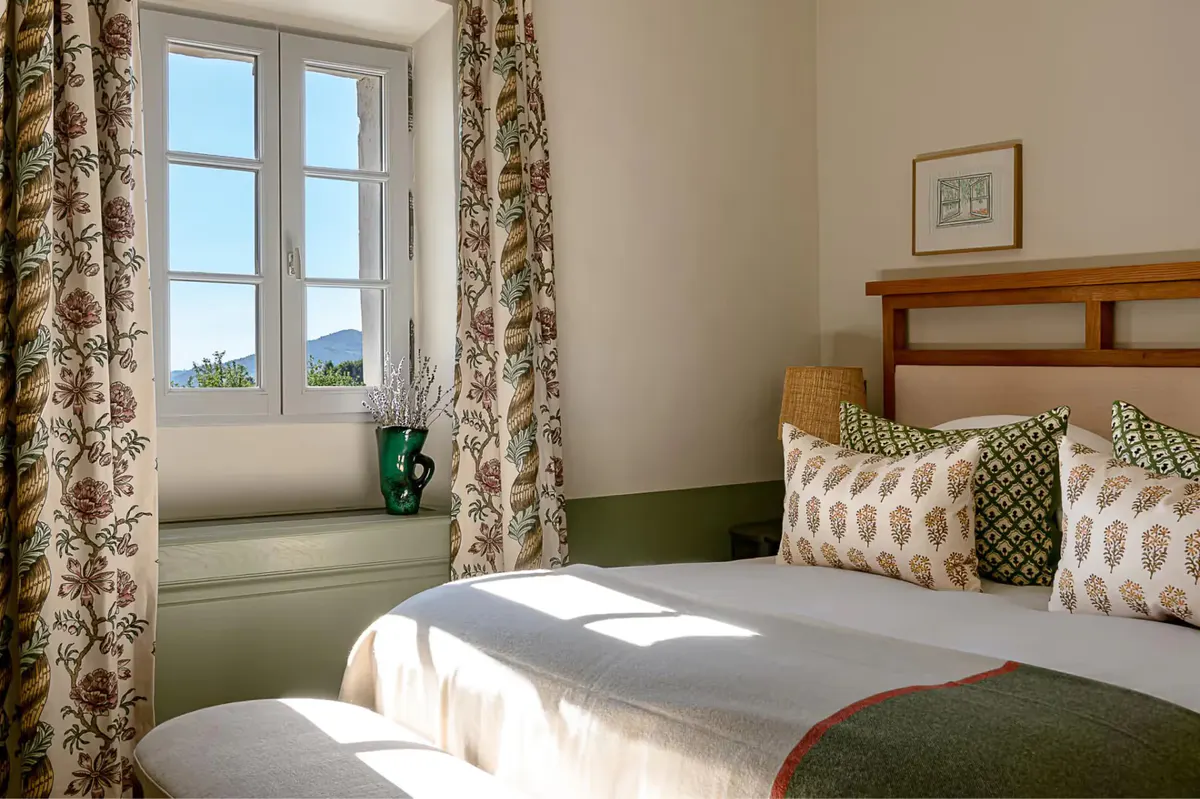 A bedroom with patterned green curtains, wooden shelving, twin beds with green blankets, and morning light