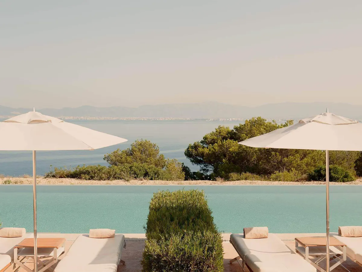 A saltwater pool edged with cream loungers and white parasols, overlooking calm turquoise bay