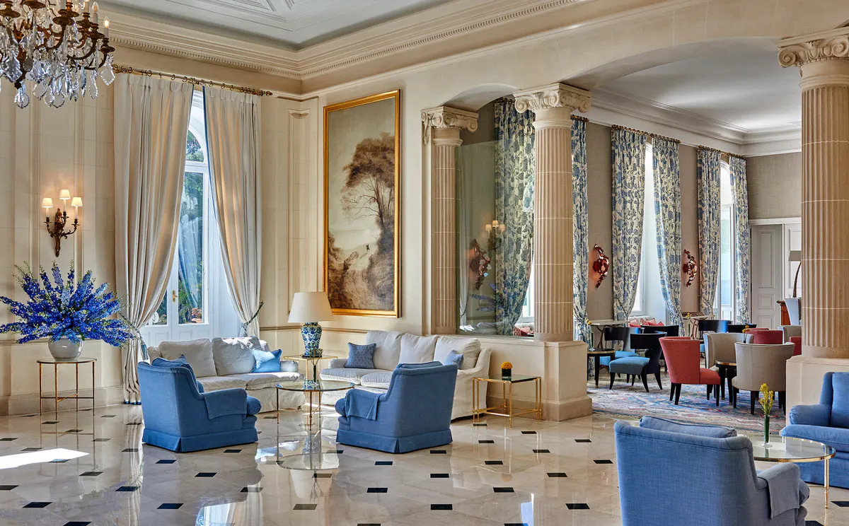 An ornate lounge with blue accents, gold leaf details, arched niches, and windows to the grounds